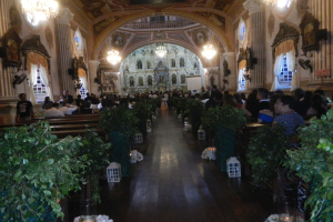 Eine Hochzeitszeremonie in einer Kirche mit sitzenden und stehenden Gästen, Topfpflanzen, die den Gang säumen, skulptierten Wänden, Vorhangfenstern, Kronleuchtern und einem Bildschirm im Hintergrund.