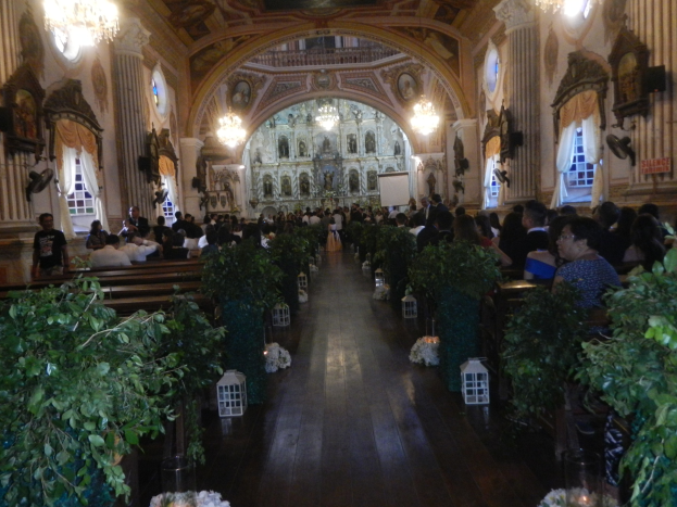 Eine Hochzeitszeremonie in einer Kirche mit sitzenden und stehenden Gästen, Topfpflanzen, die den Gang säumen, skulptierten Wänden, Vorhangfenstern, Kronleuchtern und einem Bildschirm im Hintergrund.