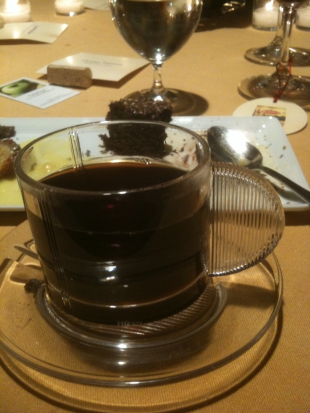 Ein Tisch mit Tasse, Untertasse, Tee, Teller, Löffel, Brownie, Wein, Kerze und weiteres Essen.