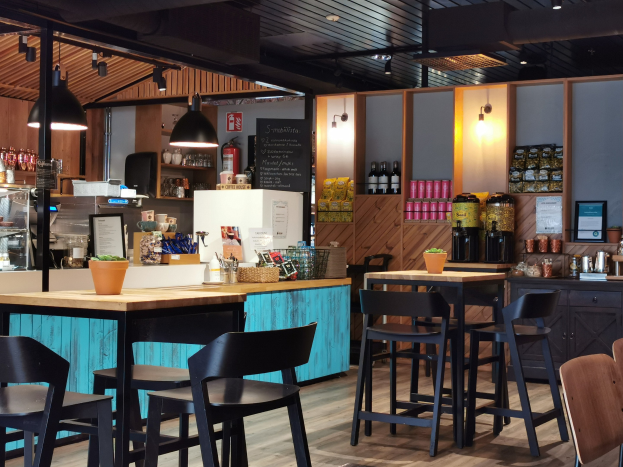 Innenraum eines Restaurants mit Holztischen, Stühlen, einer Topfpflanze, einer Schiefertafel, einem Kühlschrank, einem Feuerlöscher, Regalen mit Flaschen, Schränken mit Gegenständen, einem gerahmten Foto an der Wand und Deckenleuchten.