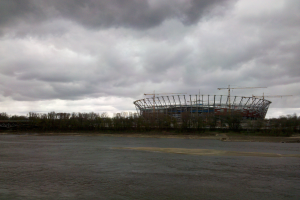 Das Olympiastadion in Kiew, Ukraine, umgeben von Bäumen und einer Brücke, mit einem bewölkten Himmel und sichtbarem Boden unten.