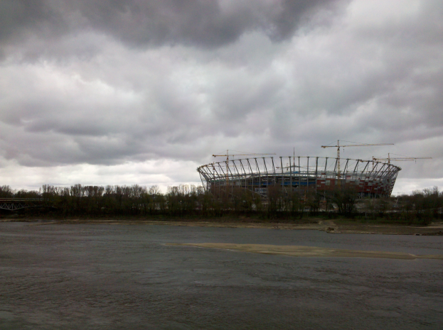 Das Olympiastadion in Kiew, Ukraine, umgeben von Bäumen und einer Brücke, mit einem bewölkten Himmel und sichtbarem Boden unten.