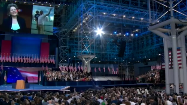 Eine große Menschenmenge vor einer Bühne mit einem Rednerpult, Mikrofon, Flaggen, einem Bildschirm und Beleuchtung, die auf eine Präsidentenrede warten.