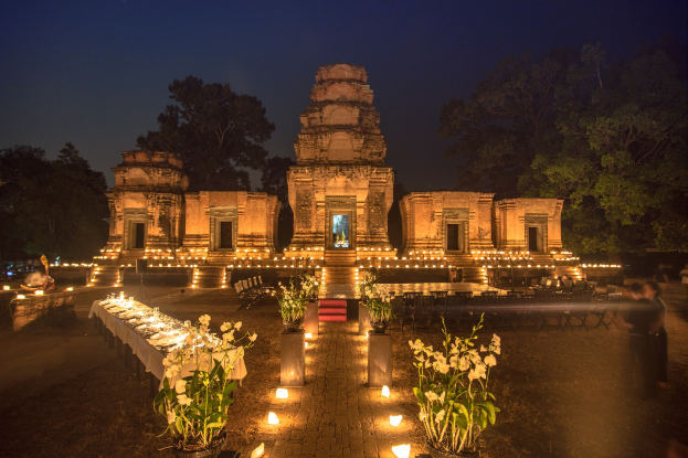 Eine Gruppe von Menschen versammelt sich nachts vor einem beleuchteten Tempel umgeben von Bäumen mit einem Tisch voller Blumentöpfe und Kerzen auf der linken Seite für eine Hochzeit in Angkor Wat.