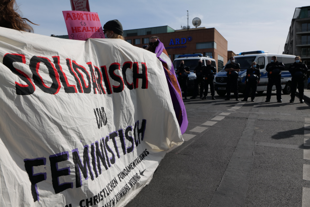 Eine Gruppe von Menschen marschiert mit einem Transparent, auf dem 'Solidarität und Feminismus' steht, an einem Gebäude vorbei, mit parkenden Fahrzeugen und Gebäuden im Hintergrund unter einem klaren blauen Himmel.