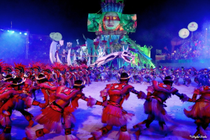 Eine Gruppe von Menschen in farbenfrohen Kostümen tanzt auf einer Bühne während des Karnevals in Rio de Janeiro 2014, umgeben von einem Publikum auf Stühlen, Lichtern, Pfählen und Ballons, und einem Bildschirm im Hintergrund, der eine Person und andere Gegenstände zeigt.