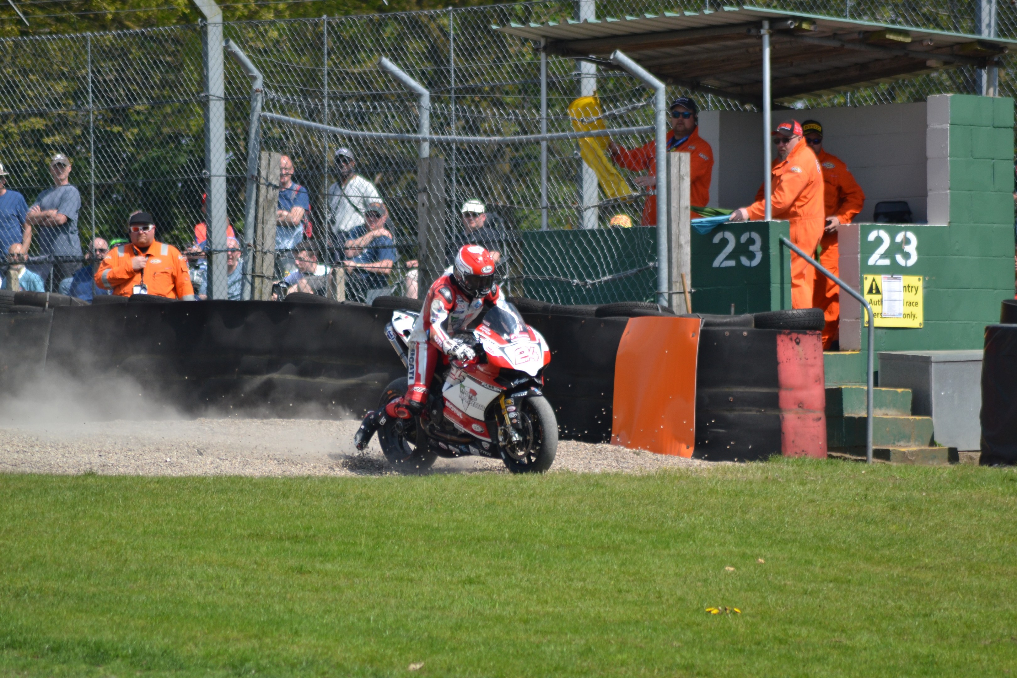 Ein Motorradfahrer mit Helm und Handschuhen fährt über ein grasbewachsenes Feld, mit Zuschauern in Mützen und Schutzbrillen, einem Zaun, Treppen mit Geländern und einem nummerierten Schuppen im Hintergrund.