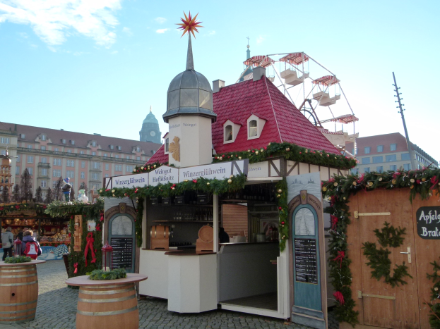 Ein kleines Gebäude mit einem roten Dach und einem Riesenrad davor, umgeben von Festdekorationen und Menschen bei einem Oktoberfest-Event in München, Deutschland.