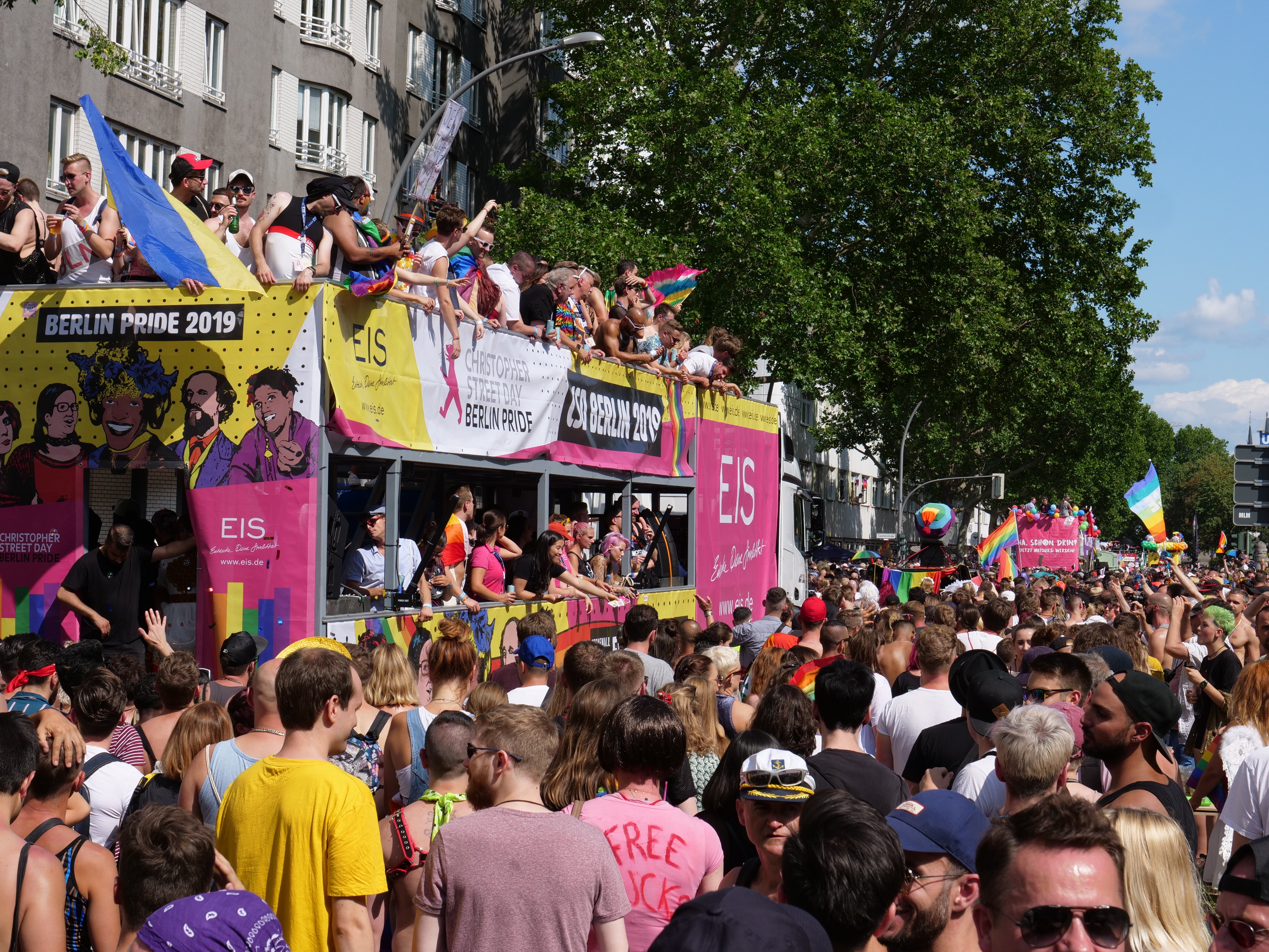 Eine große Menschenmenge marschiert auf der Straße bei der Berlin Pride 2019, viele tragen Mützen und Schutzbrillen, halten Fahnen und Schilder, mit Gebäuden im Hintergrund bei einem bewölkten Himmel.