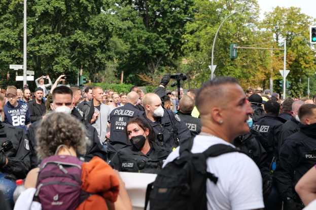 Eine große Gruppe von Menschen steht vor einer Menschenmenge von Polizisten, einige tragen Masken und Rucksäcke, andere halten Kameras, mit Laternenmasten, Verkehrsampeln, Schildern, Bäumen und einem klaren blauen Himmel im Hintergrund.