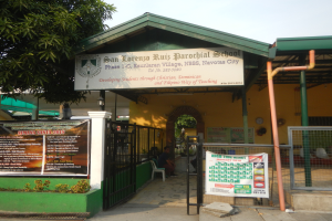 Der Eingang der San Lorenzo Ruiz Pfarrschule hat eine Tafel mit Text, einen Metallzaun, Menschen, die unter einem Dach sitzen, Pfosten, ein Schild, Pflanzen, einen Baum und einen bewölkten Himmel.
