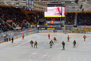 Eine Gruppe von Menschen mit Helmen und Hockeystöcken spielt ein Spiel auf einem Eisstadion mit einem Netz in der Mitte, umgeben von Texttafeln und einem großen Bildschirm im Hintergrund, wobei eine Person mit Hut zu sehen ist.