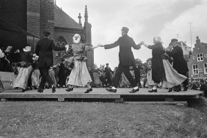 Schwarzes und wei├čes Bild einer Gruppe von Menschen, die auf einer Bühne vor einer Kirche tanzen, einige halten sich an den H├Ąnden, andere schauen zu, umgeben von Geb├Ąuden, B├Ąumen, Masten und einem klaren Himmel.
