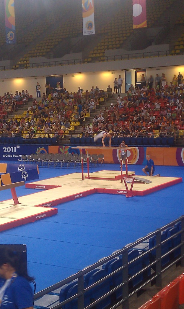 Eine Stadion-Szene mit Menschen auf Stühlen, anderen im Hintergrund, Werbetafeln oben, Treppen in der Mitte und einer Person, die in der Mitte Gymnastik betreibt.