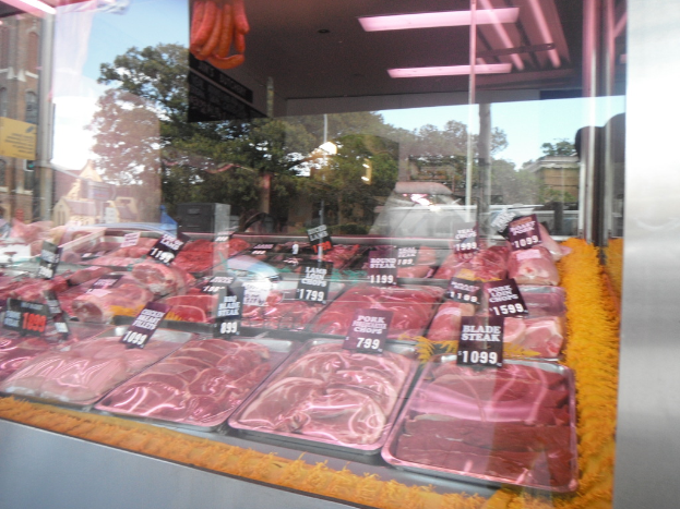 Eine Glasvitrine mit Tabletts voller Fleisch mit Preisschildern, mit Gebäuden und Bäumen durch die Glasscheibe und einer Decke oben zu sehen.