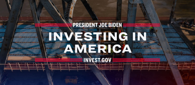 Präsident Joe Biden neben einer Brücke mit dem Text "In America investieren" und verstreuten Gegenständen im Hintergrund.