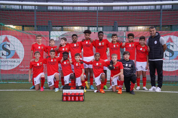 Eine Gruppe junger Männer in roten T-Shirts und schwarzen Shorts steht auf einem Fußballfeld neben einer Kiste roter Gegenstände, mit Schildern im Hintergrund, die 'FC Red Black Eglaute - Champions League' lesen.