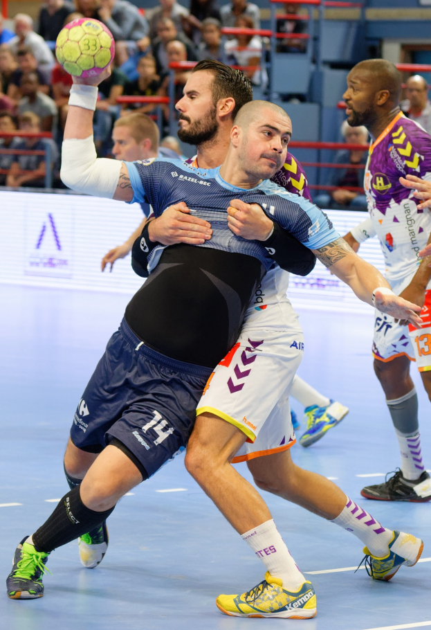 Eine Gruppe von Männern spielt Handball in einem Stadion, einer hält den Ball, mit Zuschauern im Hintergrund.