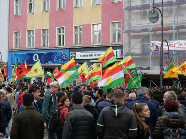 Eine große Gruppe von Menschen geht eine Straße entlang und hält kurdische Flaggen in den Händen, während Laternenmasten und Gebäude die Straße säumen und Texttafeln im Hintergrund zu sehen sind.