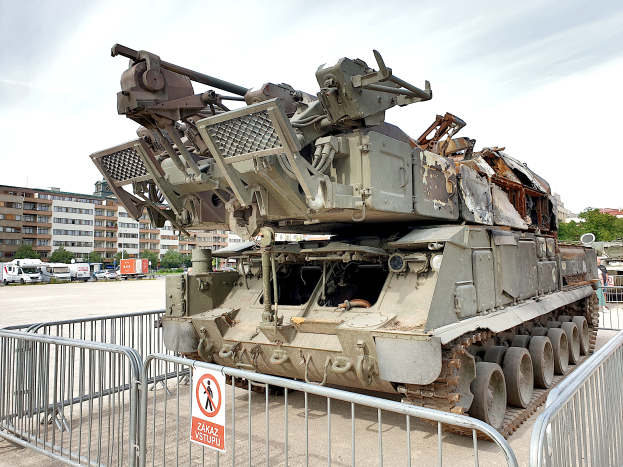Ein gepanzertes Fahrzeug vor einem Metallzaun mit einem Schild daran geparkt, mit Fahrzeugen, Bäumen, Gebäuden und einem klaren blauen Himmel im Hintergrund.