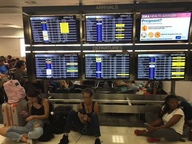 Gruppe von Menschen auf Flughafenboden sitzend, einige halten Handys, mit verstreuten Taschen und Zika-Virus-Informationen im Hintergrund.