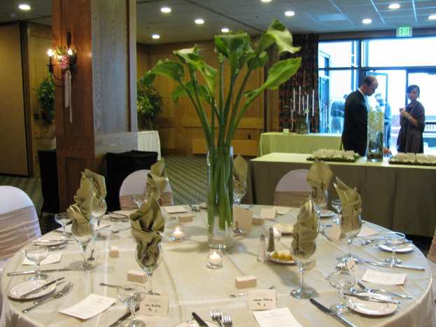 Ein Hotelrestaurant mit einer weißgedeckten Tafel, auf der Teller, Besteck, ein Glas und Zweige liegen, umgeben von Stühlen, einem grünen Tisch im Hintergrund, einem Mann in einem schwarzen Blazer, einem Mädchen neben ihm und einer Tür mit der Aufschrift "Ausgang", hinter der zwei Männer stehen.