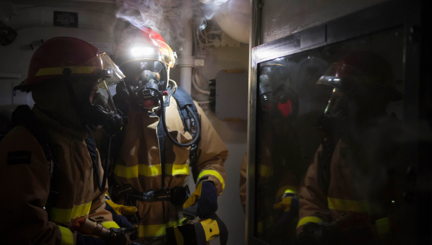 Eine Gruppe von Feuerwehrleuten in Gasmasken und Helmen steht vor einer rauchenden Tür, mit Rohren und anderen Gegenständen im Hintergrund, was auf einen begrenzten Raum hinweist.