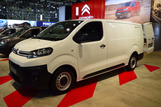 Ein weißer Citroen Berlingo Van wird auf einer Automesse präsentiert, umgeben von schwarzen Schlafzimmermöbeln, mit Wänden, die mit Brettern und Lichtern verziert sind.