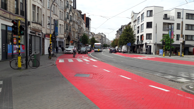 Rote Fahrradspur neben einer Stadtstraße mit fahrenden Fahrzeugen, Fußgängern auf dem Gehweg, Gebäuden mit Fenstern, Bäumen im Hintergrund und einem klaren blauen Himmel.