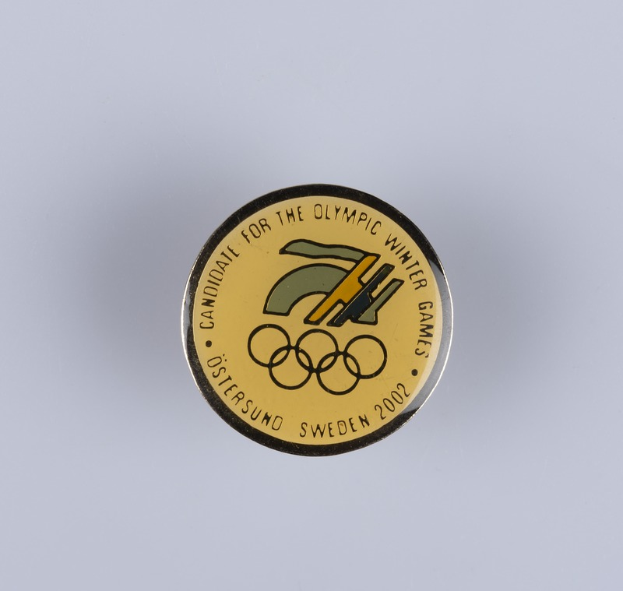 Ein runder Abzeichen mit schwarzem Hintergrund und goldfarbenem Olympia-Winterspielen-Logo in der Mitte, mit "Olympic Winter Games" in fetter weißer Schrift.