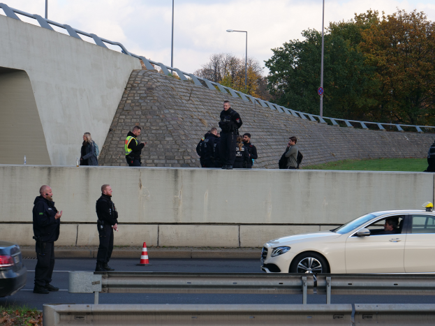 Eine Gruppe von Polizeibeamten steht neben einem Auto auf der Straße, mit Verkehrskegeln, einer Barriere, Gras, einer Wand, Laternen, Bäumen und einem bewölkten Himmel im Hintergrund.