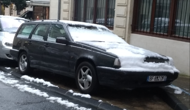 Ein schneebedecktes Auto steht am Straßenrand, mit Gebäuden und Bäumen im Hintergrund, das einen sichtbaren Reifen und Rad zeigt.