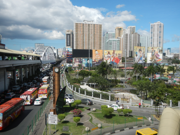 Eine belebte Stadtstraße mit zahlreichen Fahrzeugen, darunter Busse und Autos, umgeben von Gebäuden, Bäumen und einer Brücke unter einem bewölkten Himmel.