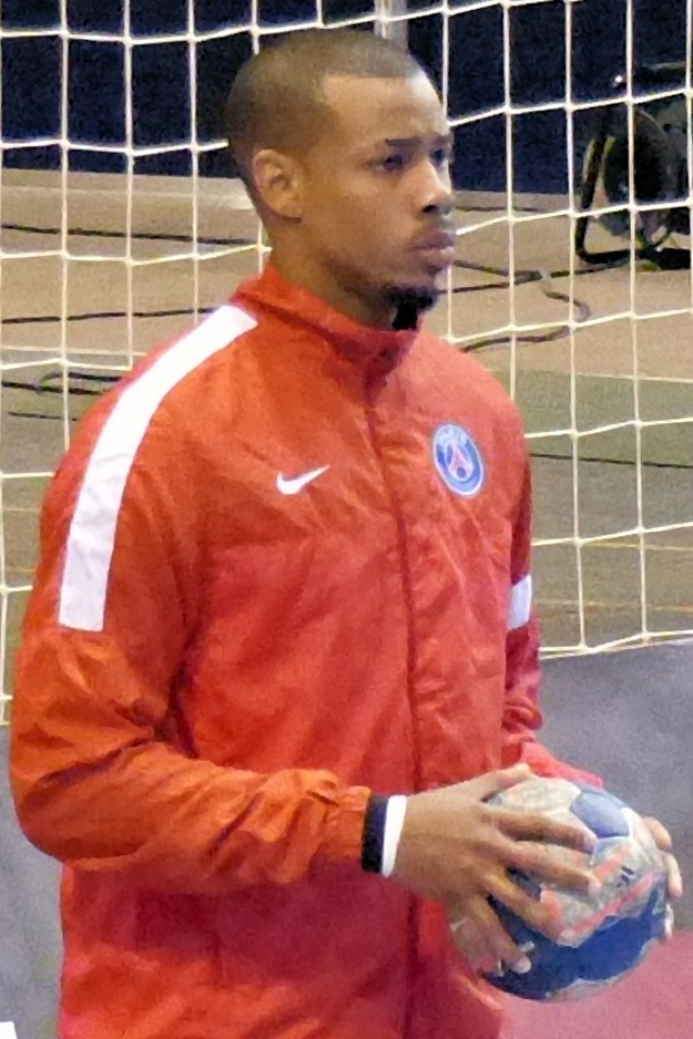 Ein Mann in einem blauen Hemd und Shorts steht vor einem Tor und hält einen Fußball mit entschlossener Miene, seine kurzen dunklen Haare und konzentrierten Augen auf den Ball gerichtet.