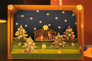 Eine hölzerne Krippe in einer Schachtel auf einem Tisch mit einer Lampe links und einer Wand im Hintergrund, dekoriert mit Weihnachtsbäumen, Tieren und einem Haus.