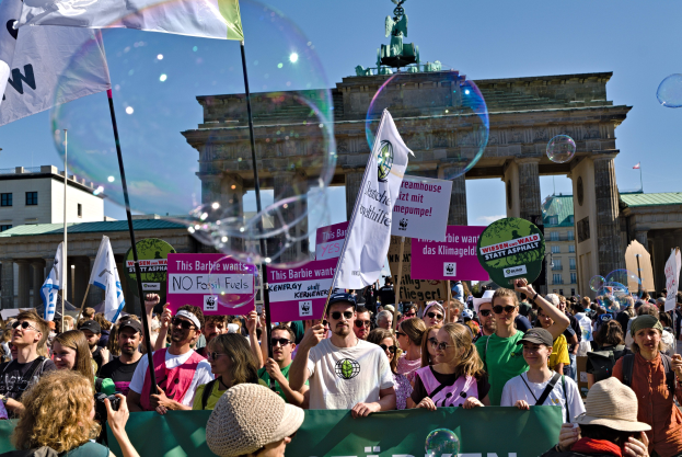 Menschenmenge vor dem Brandenburger Tor in Berlin, viele mit Mützen und Brillen, einige mit Fahnen und Spruchbändern, im Hintergrund Gebäude, ein statuegekrönter Bogen und ein mit Seifenblasen gefüllter Himmel.