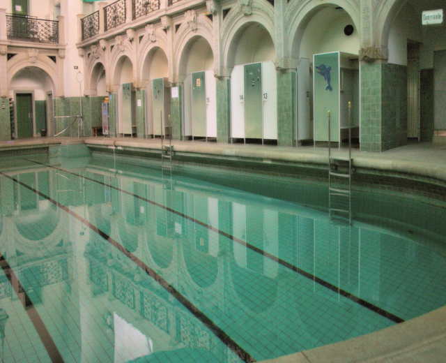 Innenpool mit klarem Wasser, umgeben von gefliestem Boden, Bogengängen, Säulen, Geländern, Türen und Texttafeln in einem große Gebäude.