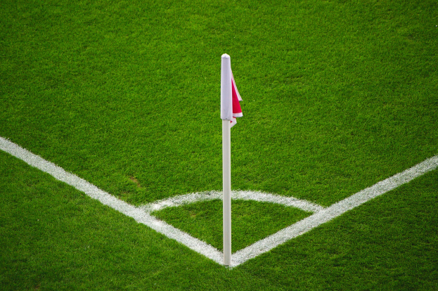 Ein Fußballfeld mit grünem Rasen und weißen Begrenzungslinien, mit einem weißen Pfosten mit einer roten und weißen Fahne in der Mitte des Strafraums.