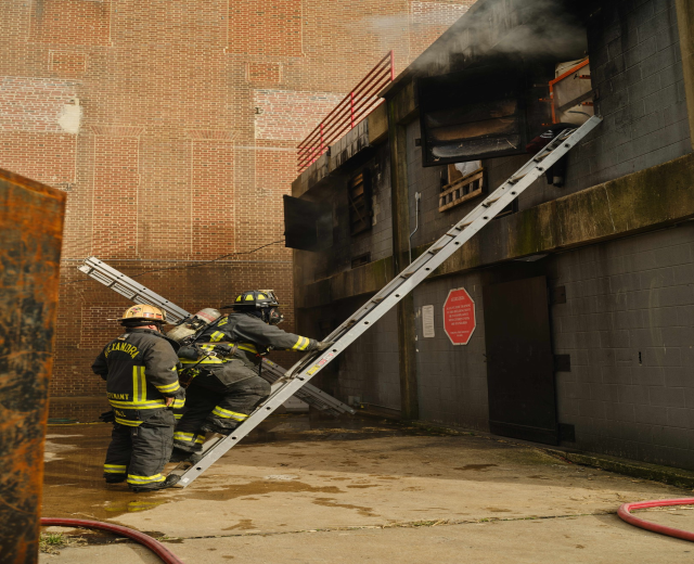 Feuerwehrleute in Helmen klettern an einer Leiter zu einem rauchgefüllten Gebäude, mit einem Metallobjekt auf der linken Seite und Rohren an der Basis.