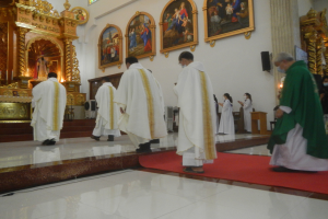 Eine Gruppe von Priestern geht eine rote Teppich in einer Kirche hinunter, mit Menschen, die auf dem Boden vor und rechts von ihnen stehen, und Rahmen an der Hintergrundwand.