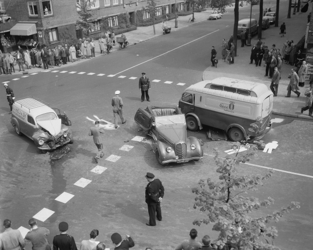 Eine Schwarz-Weiß-Szene eines Autounfalls an einer Straßenecke, mit mehreren Fahrzeugen, darunter einem Lieferwagen, einer Gruppe von Menschen drumherum und im Hintergrund Gebäude mit Fenstern, Bäumen, Masten und Texttafeln.