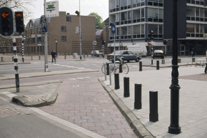 Eine Stadtstraße mit einem geparkten Fahrrad, fahrenden Fahrzeugen, Fußgängern, Pfählen, Verkehrsampeln, Schildern, Bäumen, Gebäuden mit Fenstern und einem klaren blauen Himmel.