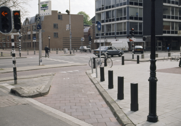 Eine Stadtstraße mit einem geparkten Fahrrad, fahrenden Fahrzeugen, Fußgängern, Pfählen, Verkehrsampeln, Schildern, Bäumen, Gebäuden mit Fenstern und einem klaren blauen Himmel.