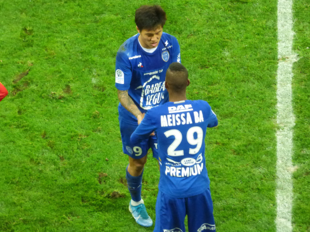 Zwei Fußballspieler in blauen Trikots stehen auf einem grünen Feld, mit einer Person in rotem Shirt links daneben.