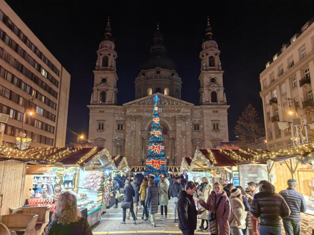 Ein geschmückter Weihnachtsmarkt mit Ständen, Menschen und festlicher Beleuchtung vor einer Kirche bei Nacht, vor dem Hintergrund von Gebäuden, Bäumen und einem sternenklaren Himmel.