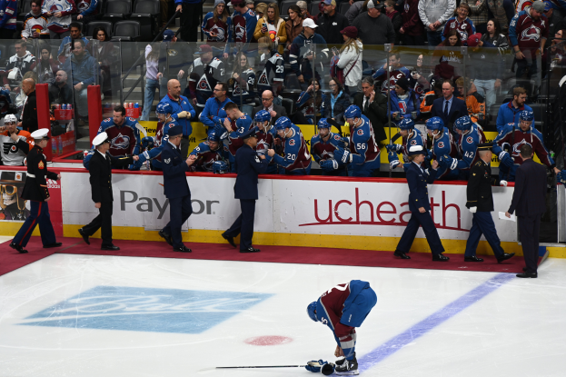 Ein Hockey-Spieler auf dem Eis mit einem Stock in der Hand, umgeben von Menschen mit Mützen und Hockey-Stöcken, mit Zuschauern und Gittern mit der Aufschrift "Colorado Avalanche vs San Jose Sharks - 3/24/15 NHL Pick, Odds, and Prediction" im Hintergrund.