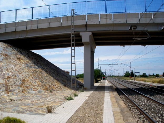 Ein Bahnhof mit einer Steinmauer und einer Brücke darüber, mit Geländern, Säulen, Strommasten mit Drähten, einer Leiter zum Bahnsteig und Bäumen mit bewölktem Himmel im Hintergrund.