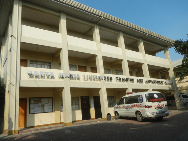 ├übersicht des Santa Maria Lebensmittel-Trainings- und Arbeitszentrums mit Fenstern, Türen und Beschriftung, umgeben von B├Ąumen und Fahrzeugen auf einer Stra├če unter einem klaren blauen Himmel.