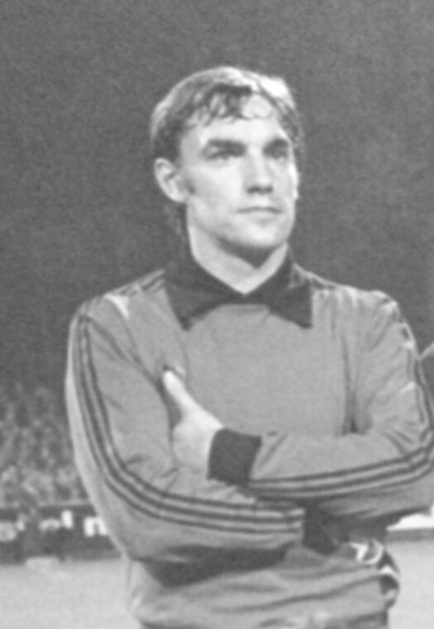 Ein Schwarz-Weiß-Foto eines Mannes, der mit verschränkten Armen auf einem Fußballfeld steht und von einer Gruppe von Menschen im Hintergrund umgeben ist, die als legendärer Torwart von Manchester United identifiziert wird.