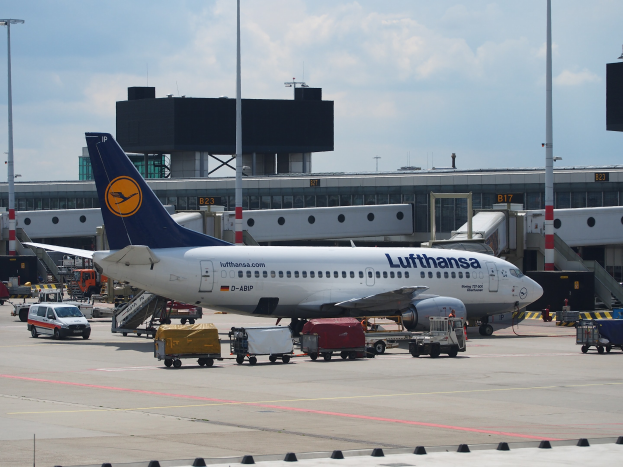 Lufthansa Airbus A320-200-Flugzeug am Frankfurter Flughafen geparkt mit Bodenfahrzeugen und Wagen im Vordergrund, einem Gebäude und Laternenmasten im Hintergrund unter einem klaren blauen Himmel.